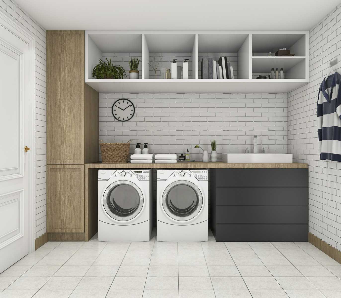 Modern laundry room with a front-load washer and dryer under a wooden countertop, surrounded by built-in cabinetry, a sink, open shelving with decor and storage items, and a white brick wall with a black clock.