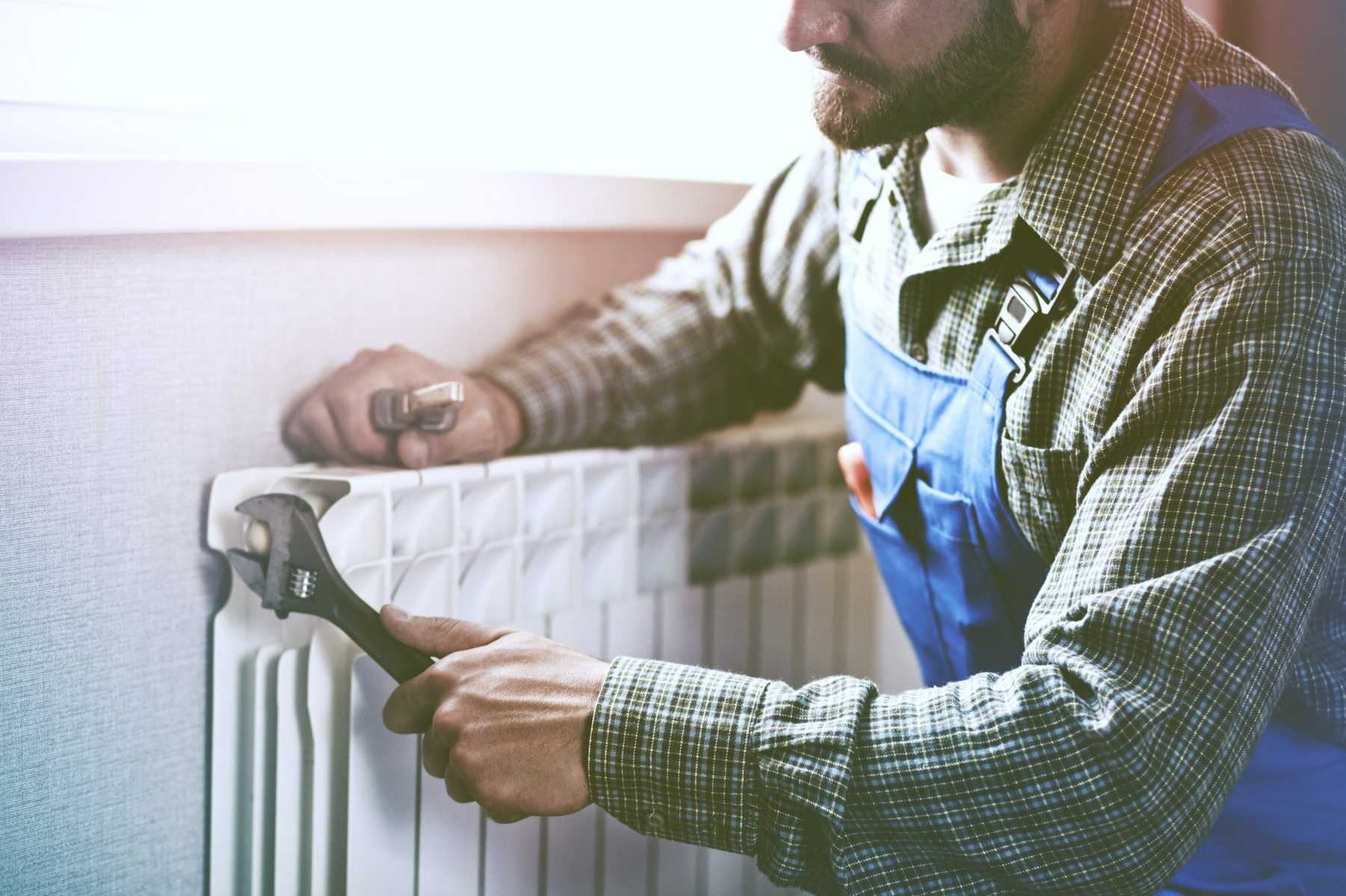 Technician in blue overalls using tools to repair or install a white radiator attached to a wall, with natural light coming through a nearby window.